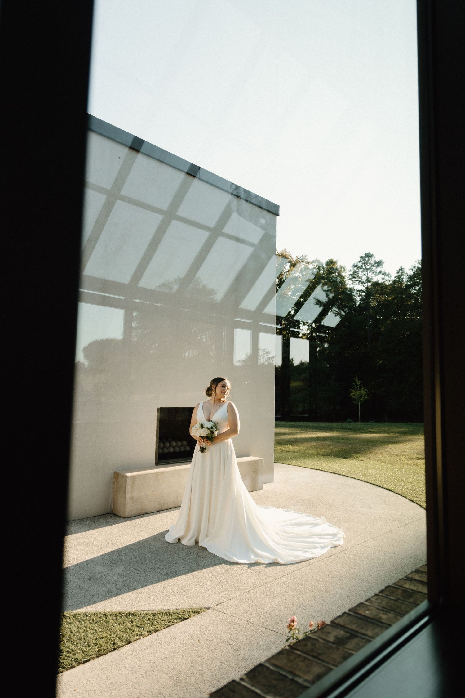 A bride in a flowing white gown with train stands in sunlight against a modern white building with glass panels.