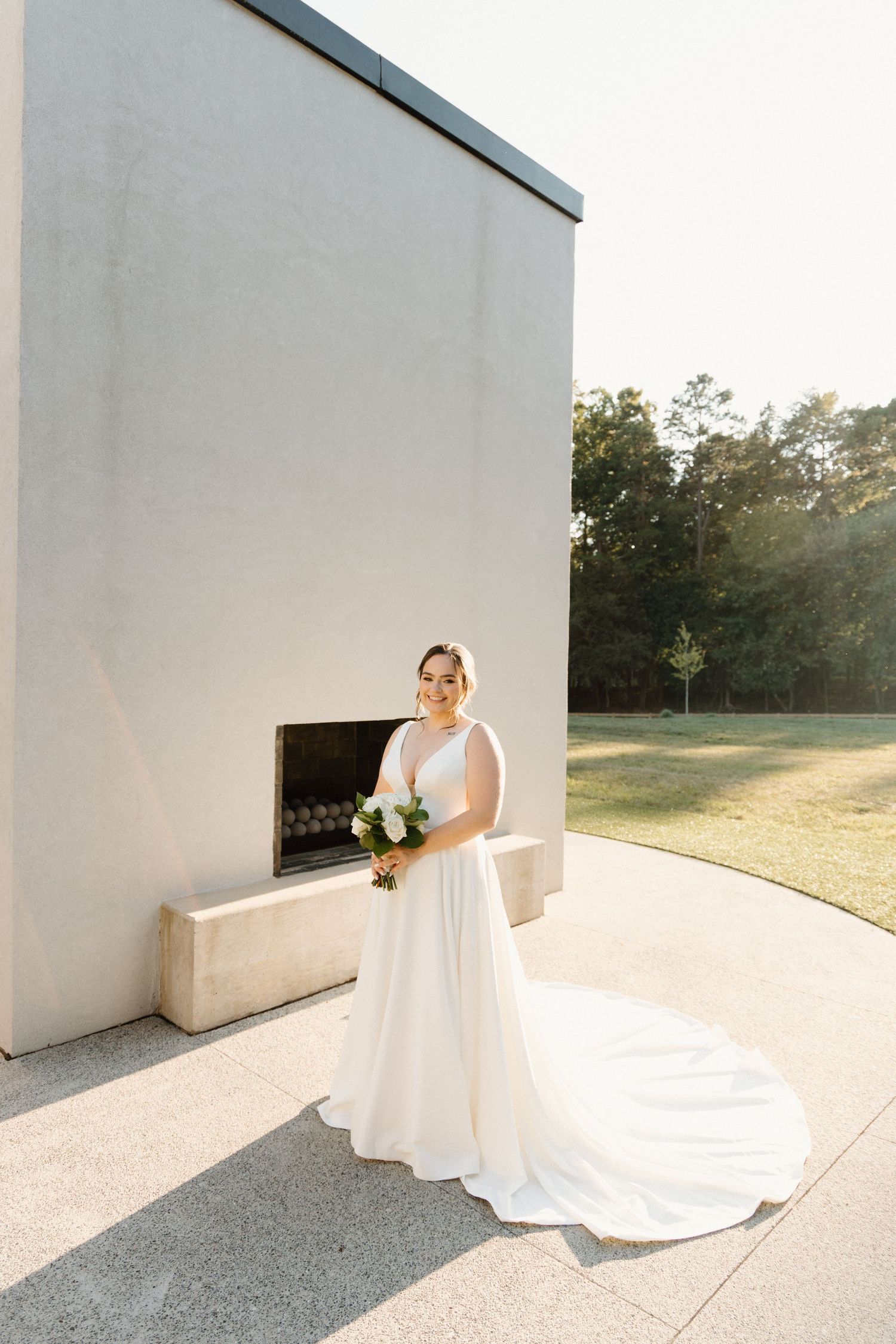 A bride in a flowing white wedding gown poses elegantly against white architectural walls in natural sunlight.