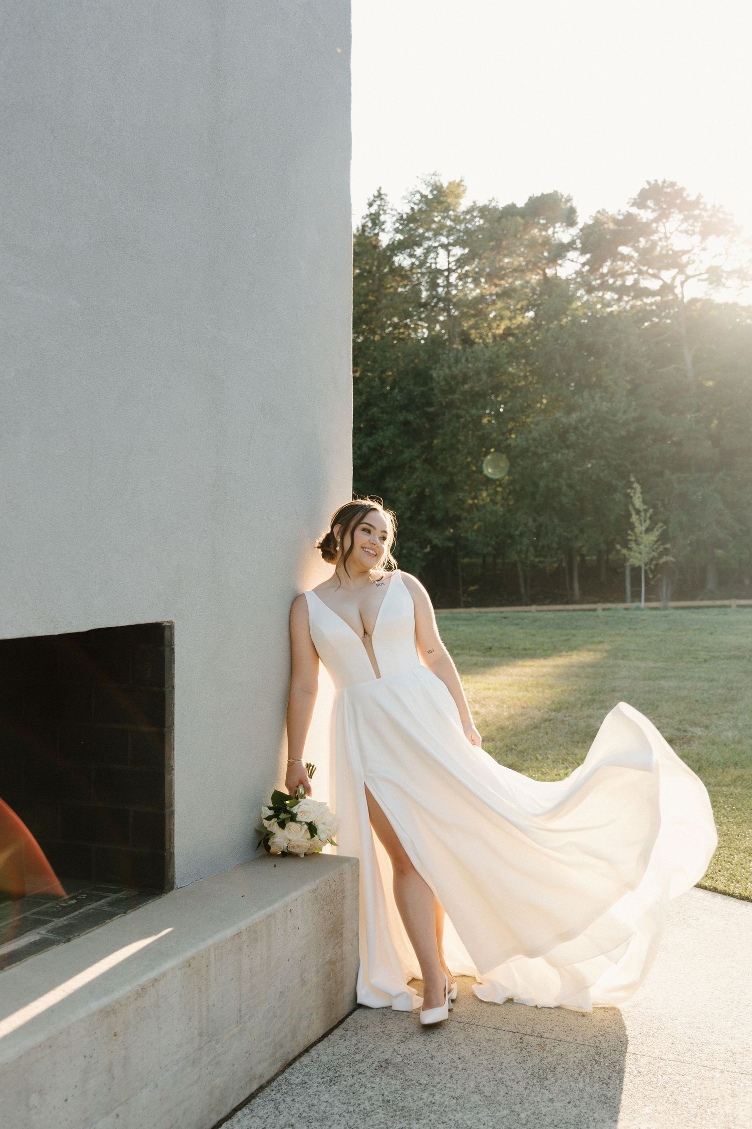 A flowing white wedding dress billows in the sunlight as someone sits on a concrete ledge in an outdoor setting.