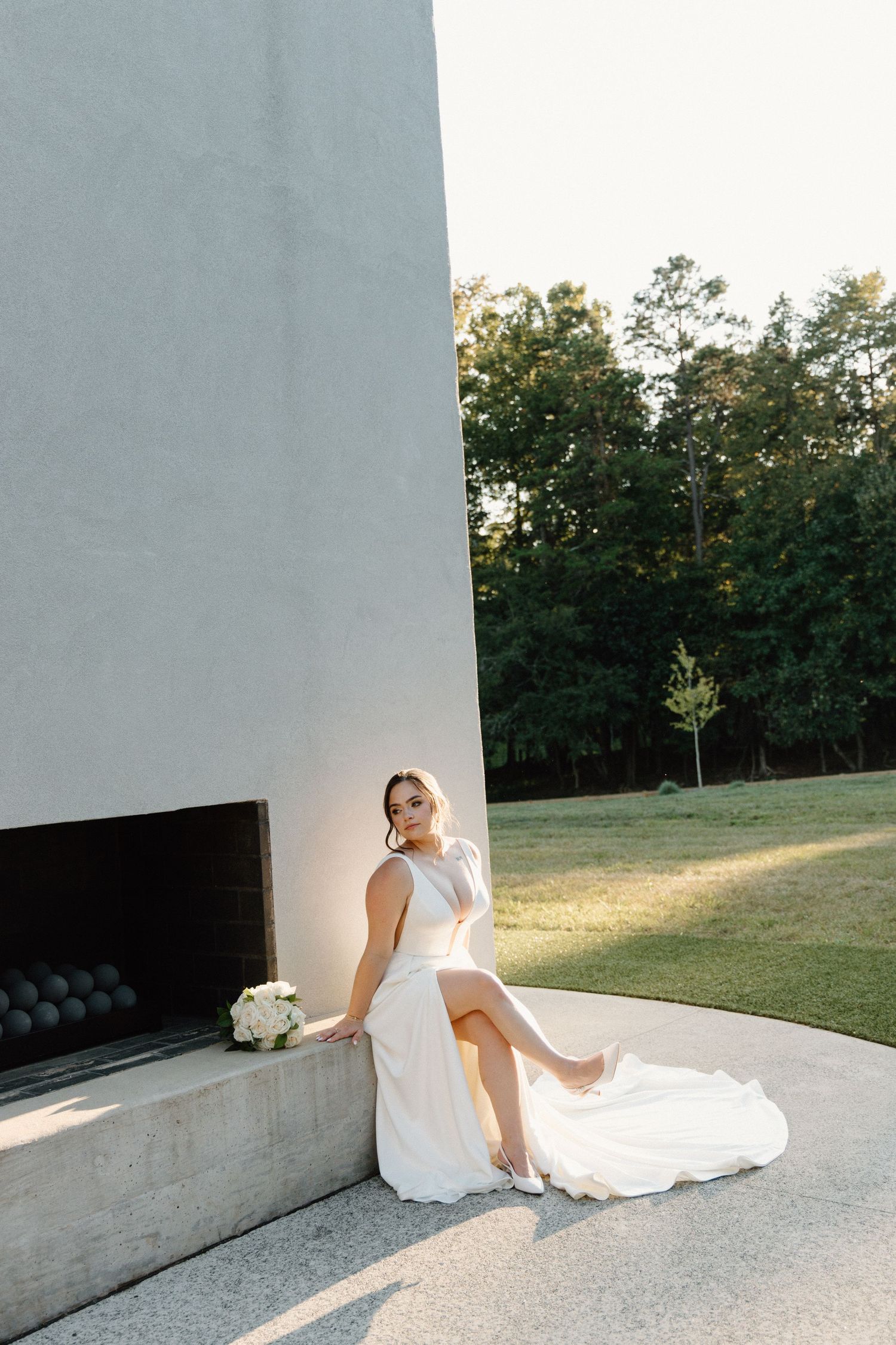 A person in a white dress sits on a concrete surface in an outdoor setting with trees in the background.