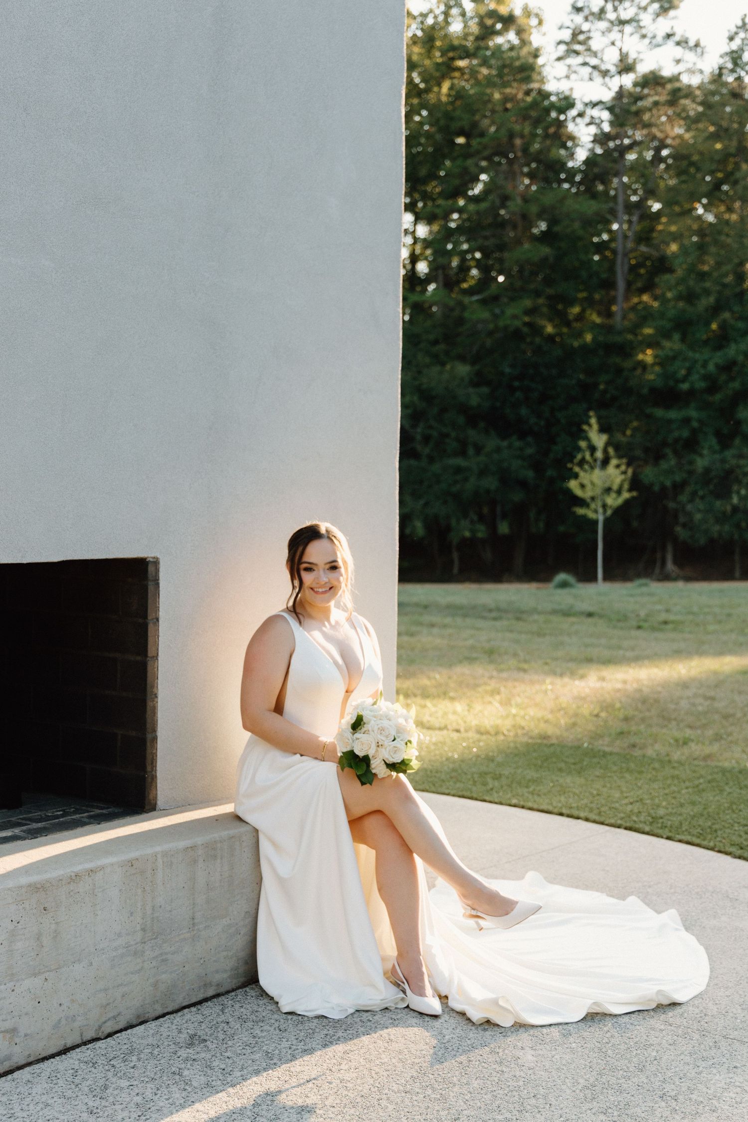 A bride in a white wedding dress sits elegantly on a modern concrete ledge in an outdoor garden setting.