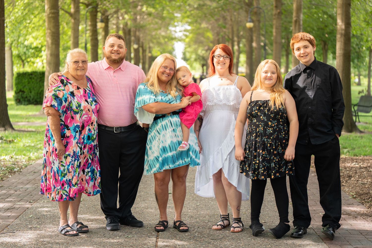 A group poses together on a tree-lined path wearing colorful dresses and formal attire.