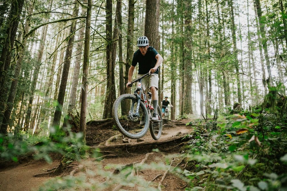 Adventurous couple engagement session photo of couple biking down trail hill in Cumberland Forest Vancouver Island doing jump