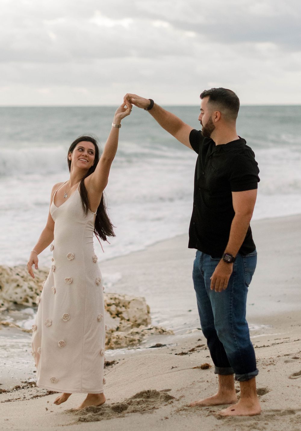 A couple dances playfully together on a sandy beach with ocean waves in the background.