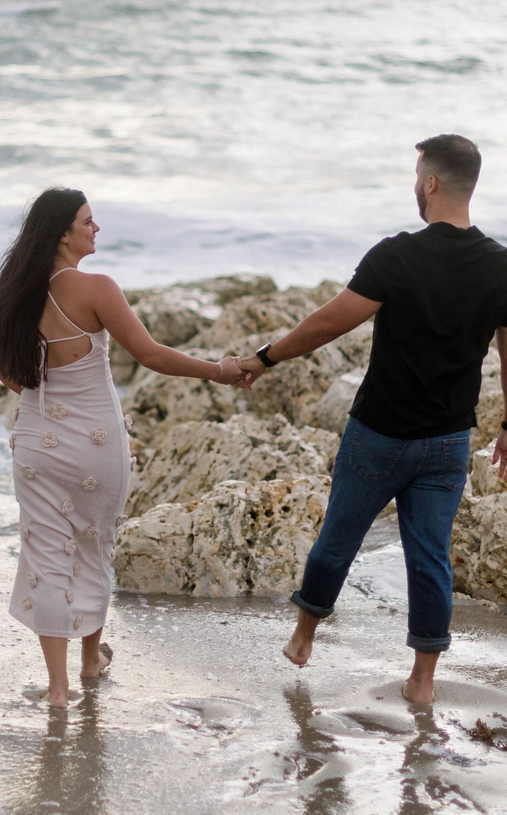 Couple walking together along rocky beach shore while waves crash against their feet in sequence of candid photos.