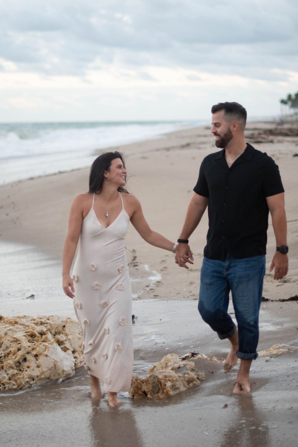 A couple walks barefoot along a cloudy beach holding hands, with the woman in a white dress and man in dark shirt and jeans.