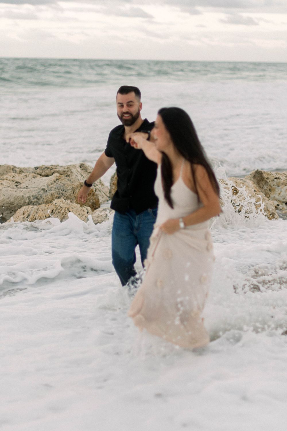 A couple playfully runs through ocean waves on a beach, with the woman's white dress flowing in the wind.