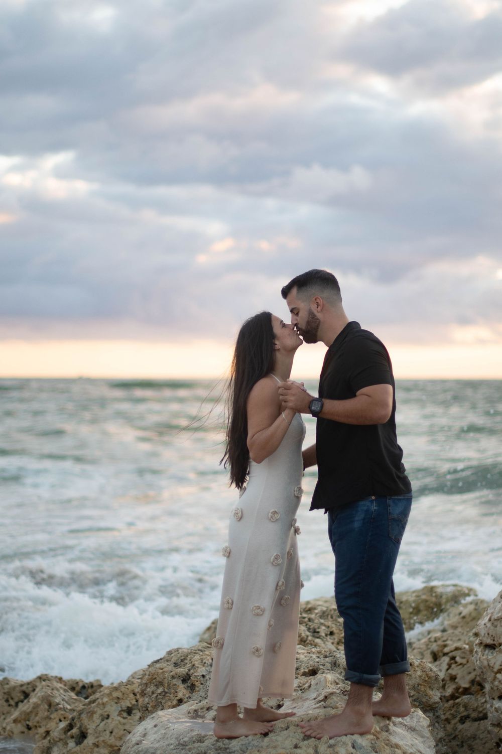 A romantic couple shares an intimate moment on rocky coastal cliffs at sunset with crashing waves in the background.