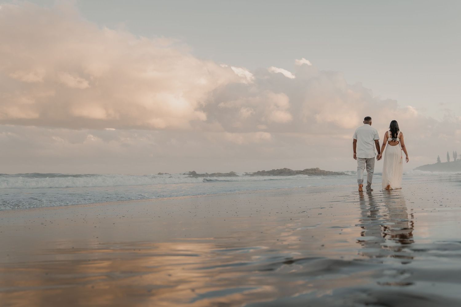 Couple walking hand in hand along reflective wet beach at sunset under dramatic cloudy sky.