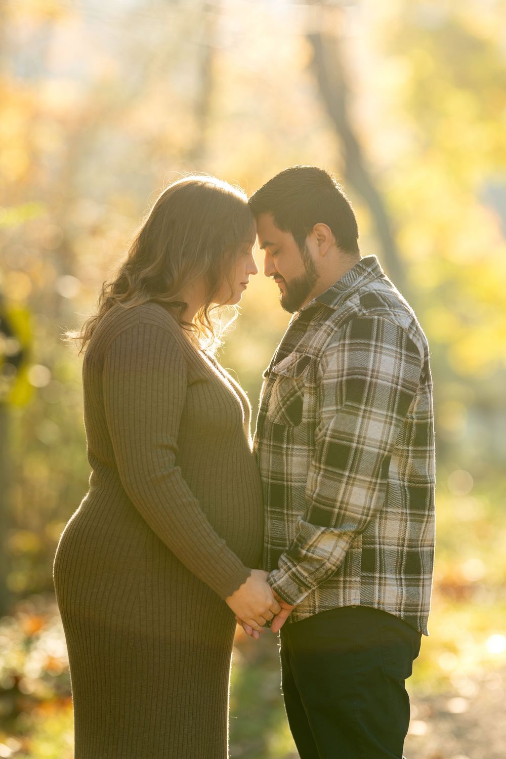 Expecting couple stands forehead-to-forehead holding hands in glowing autumn sunlight.
