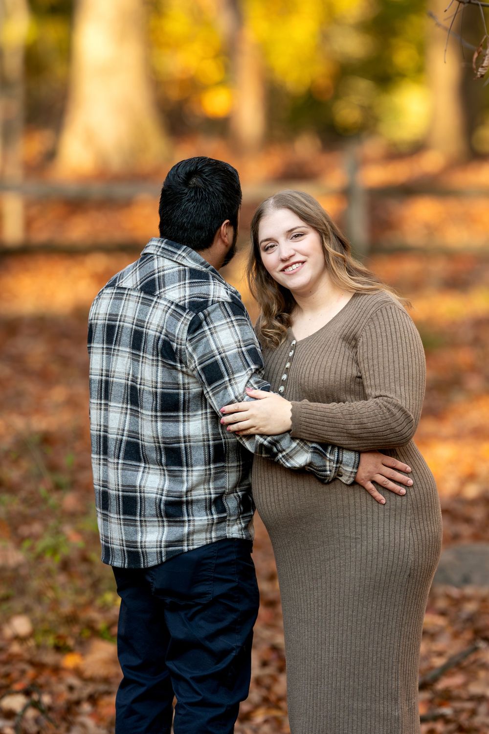 Pregnant woman smiles over shoulder at camera while partner holds her bump in a colorful autumn forest setting.