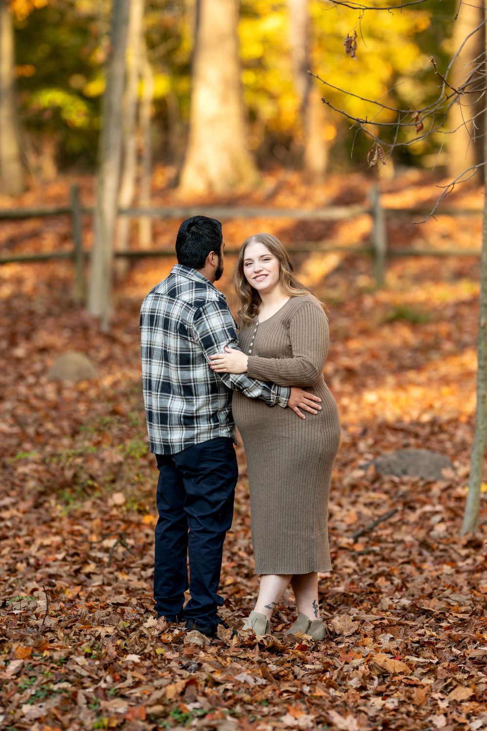 Smiling pregnant woman in brown knit dress faces partner among fallen leaves in a sunlit autumn woodland scene.