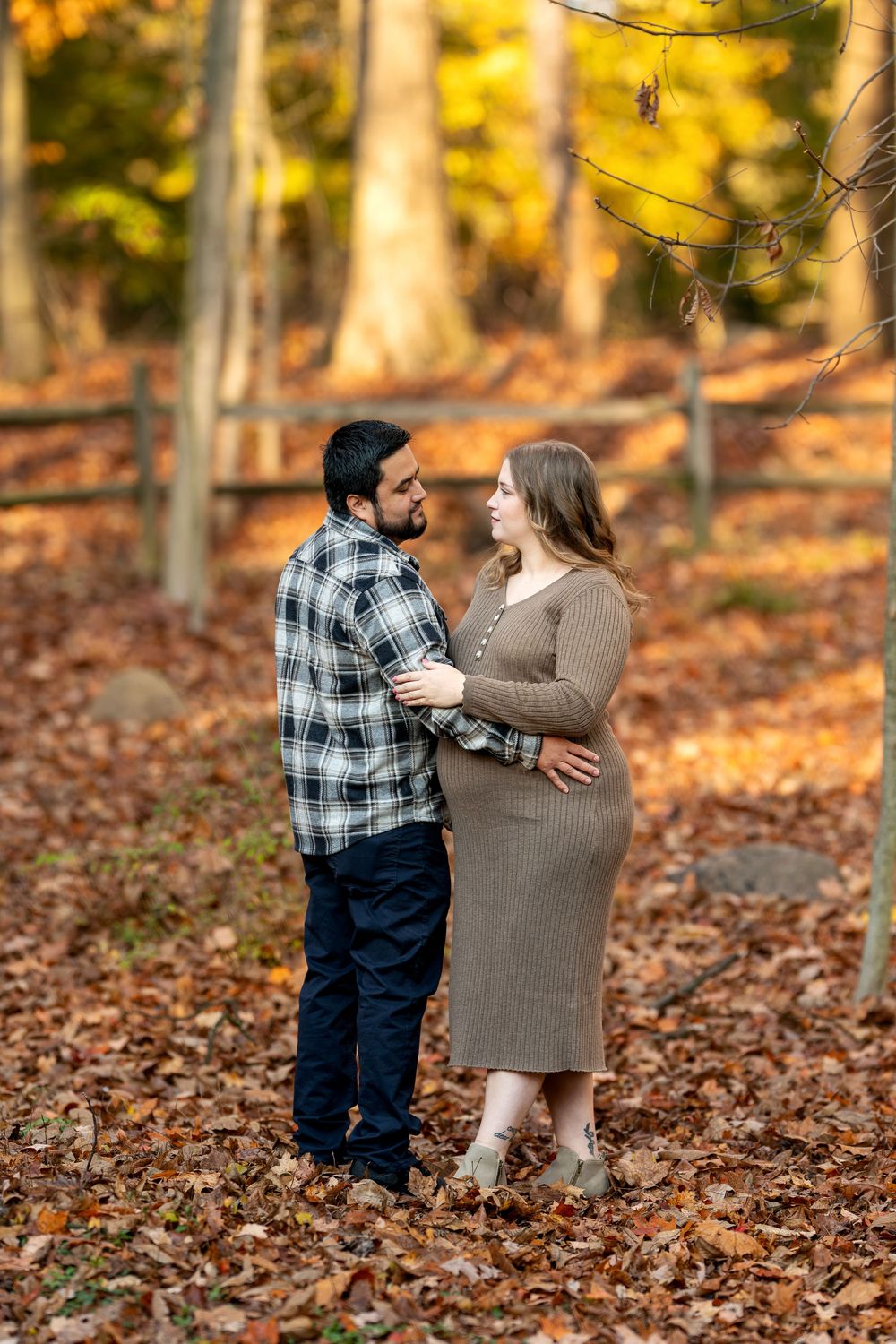 Expecting couple gazes at each other lovingly in a fall forest, surrounded by golden leaves and wooden fence.