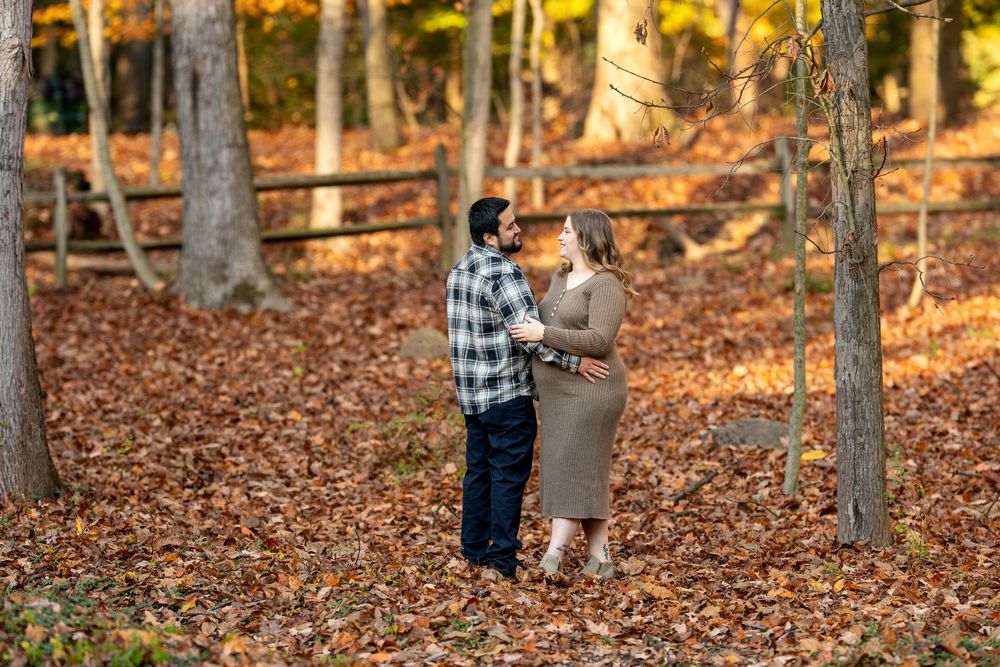 Couple stands close together in a leaf-covered autumn forest path, golden trees and wooden fence in background.