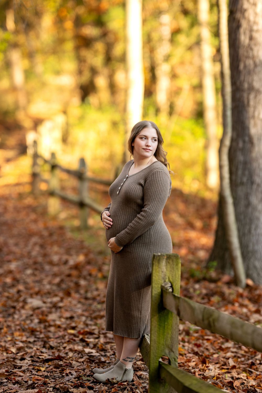 Pregnant woman in olive ribbed dress cradles baby bump beside rustic wooden fence in a sunlit autumn forest.