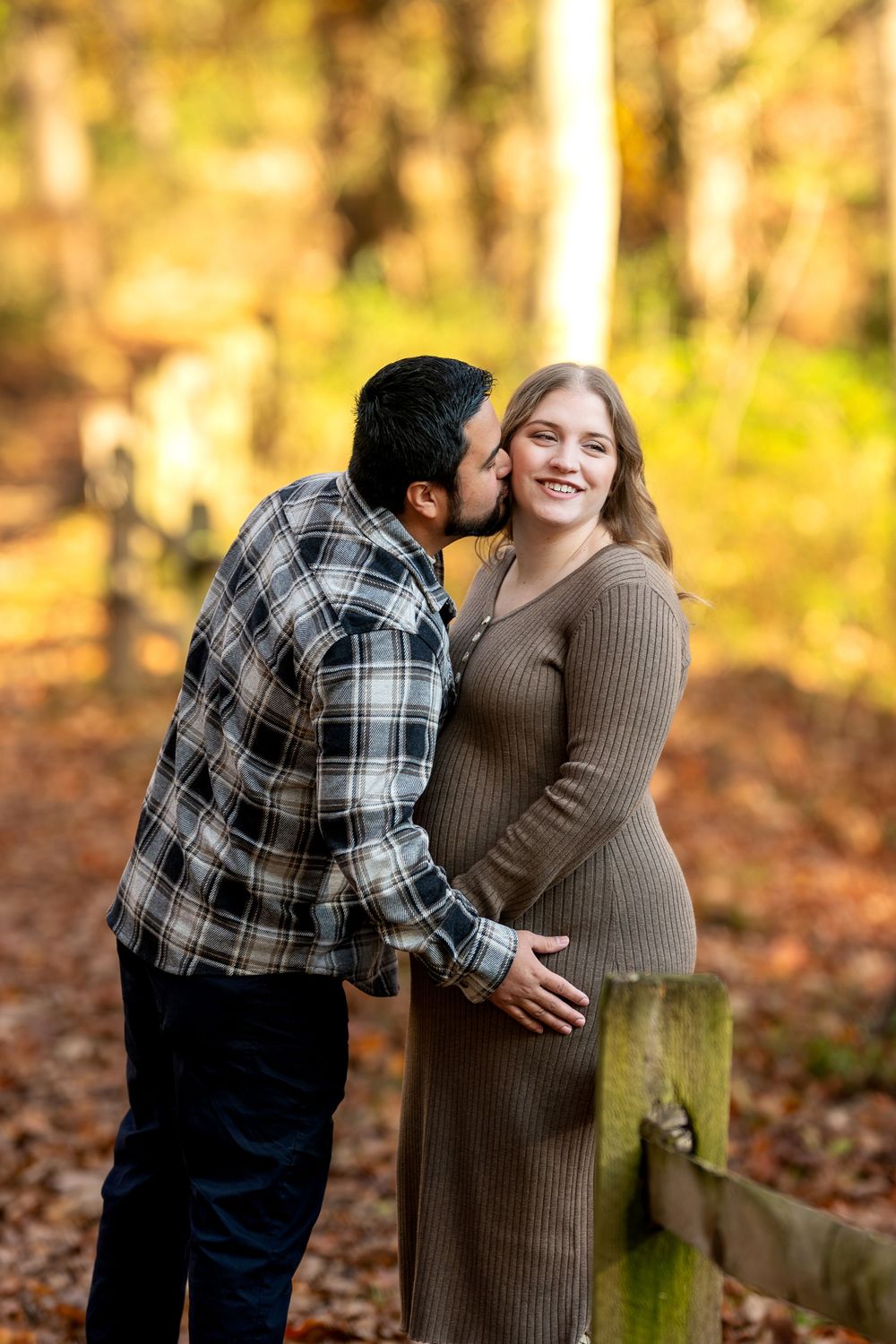 Man kisses smiling pregnant woman on cheek near wooden fence in a warm golden autumn forest setting.