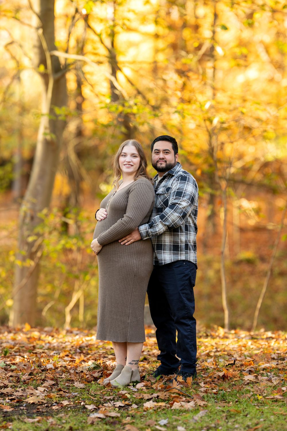 Pregnant woman in brown dress with partner embracing her from behind in fall woodland setting.