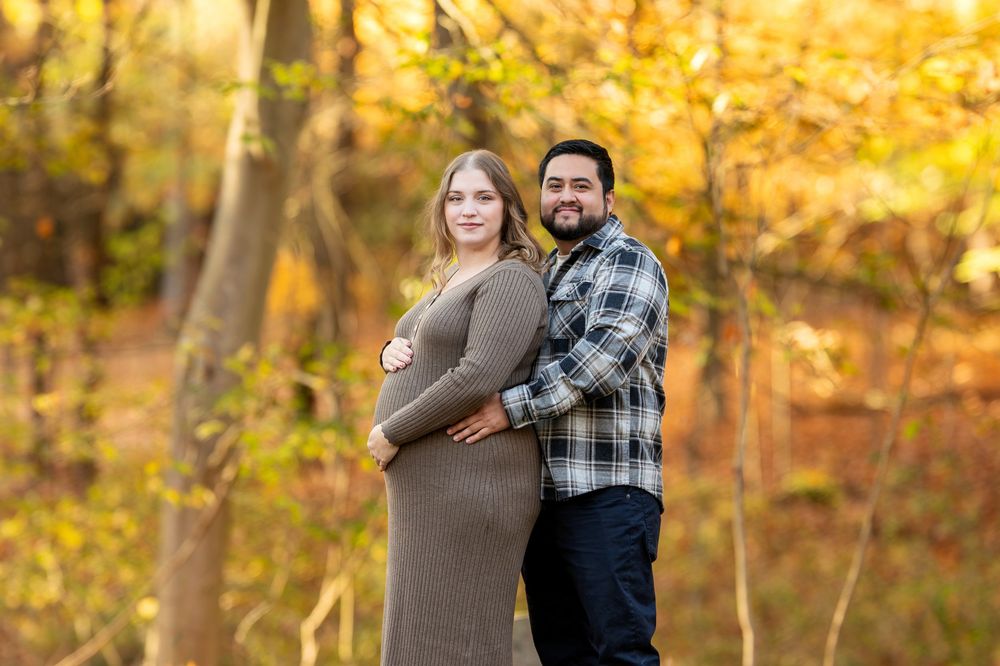 Expecting couple poses together in sunlit autumn woods, woman in brown knit dress cradling baby bump, man in plaid.