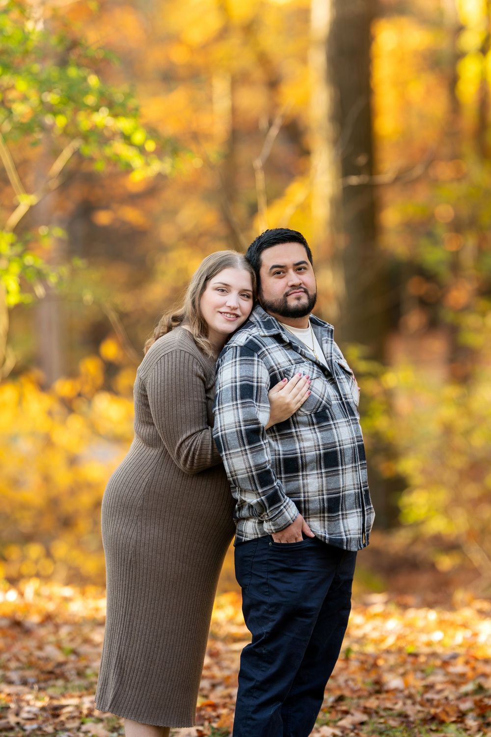 Couple embracing in autumn forest, woman in brown dress hugging man in plaid shirt among golden leaves.