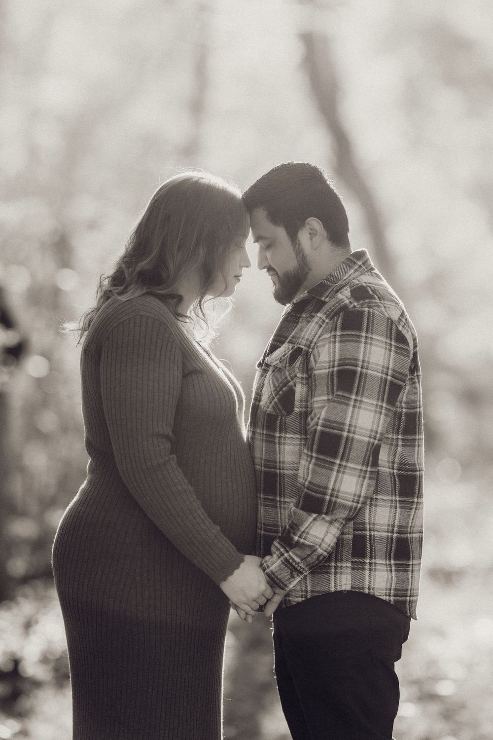Black and white maternity photo of couple holding hands and touching foreheads in a serene forest.
