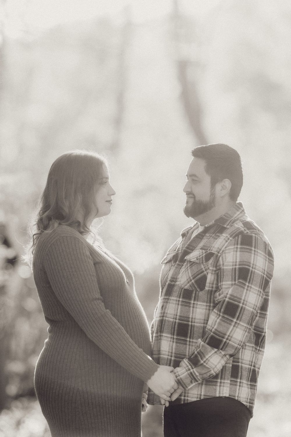 Black and white photo of a pregnant couple holding hands and gazing at each other outdoors in winter.