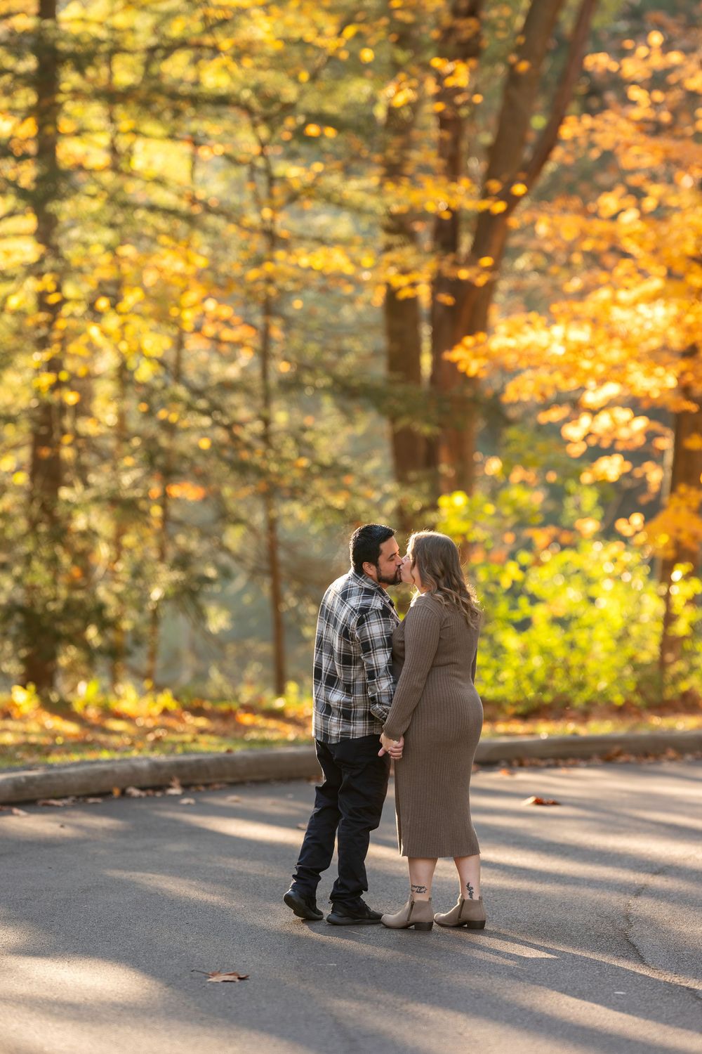 Pregnant couple shares a kiss while holding hands on a sunlit autumn road.