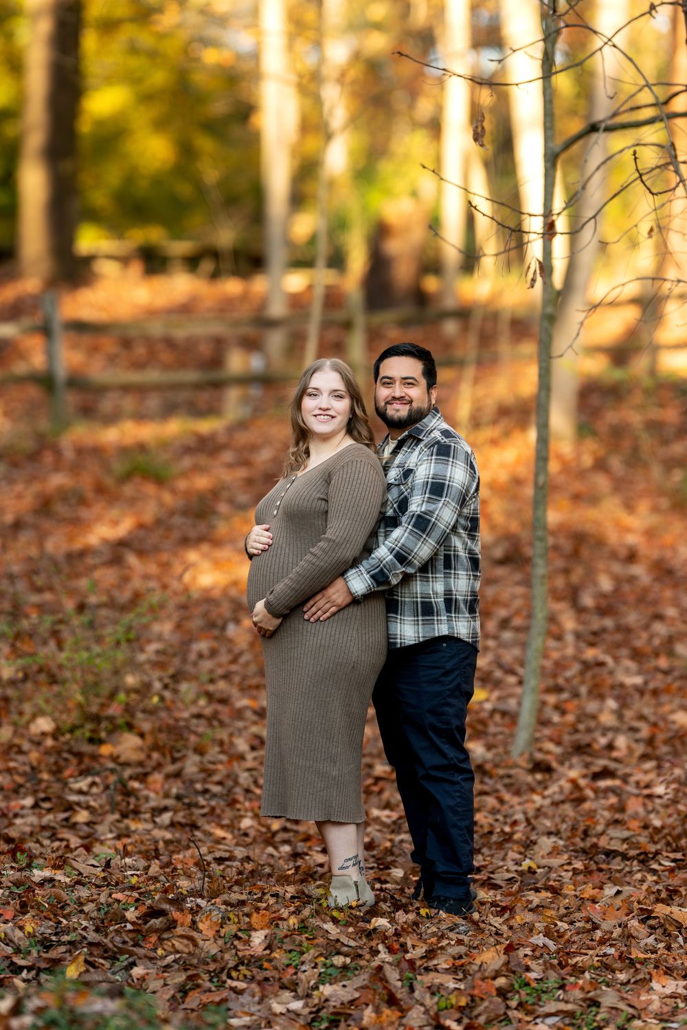 Couple poses back-to-back in autumn forest, pregnant woman in brown knit dress, man in plaid shirt, fall foliage background.