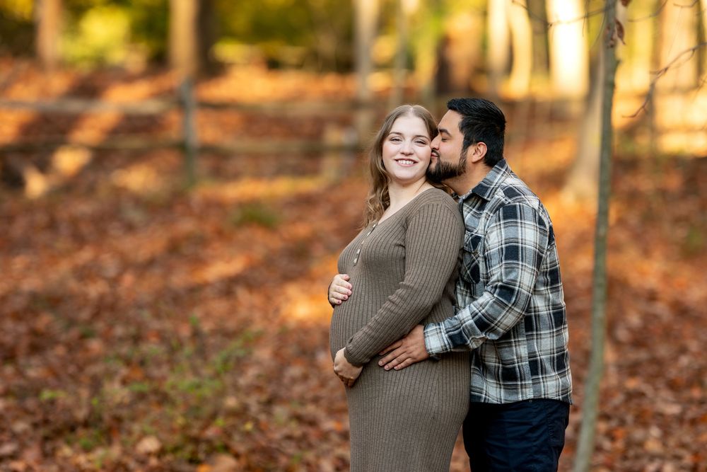 Man kisses pregnant woman's cheek tenderly in golden autumn woodland setting, she wears a ribbed brown maternity dress.