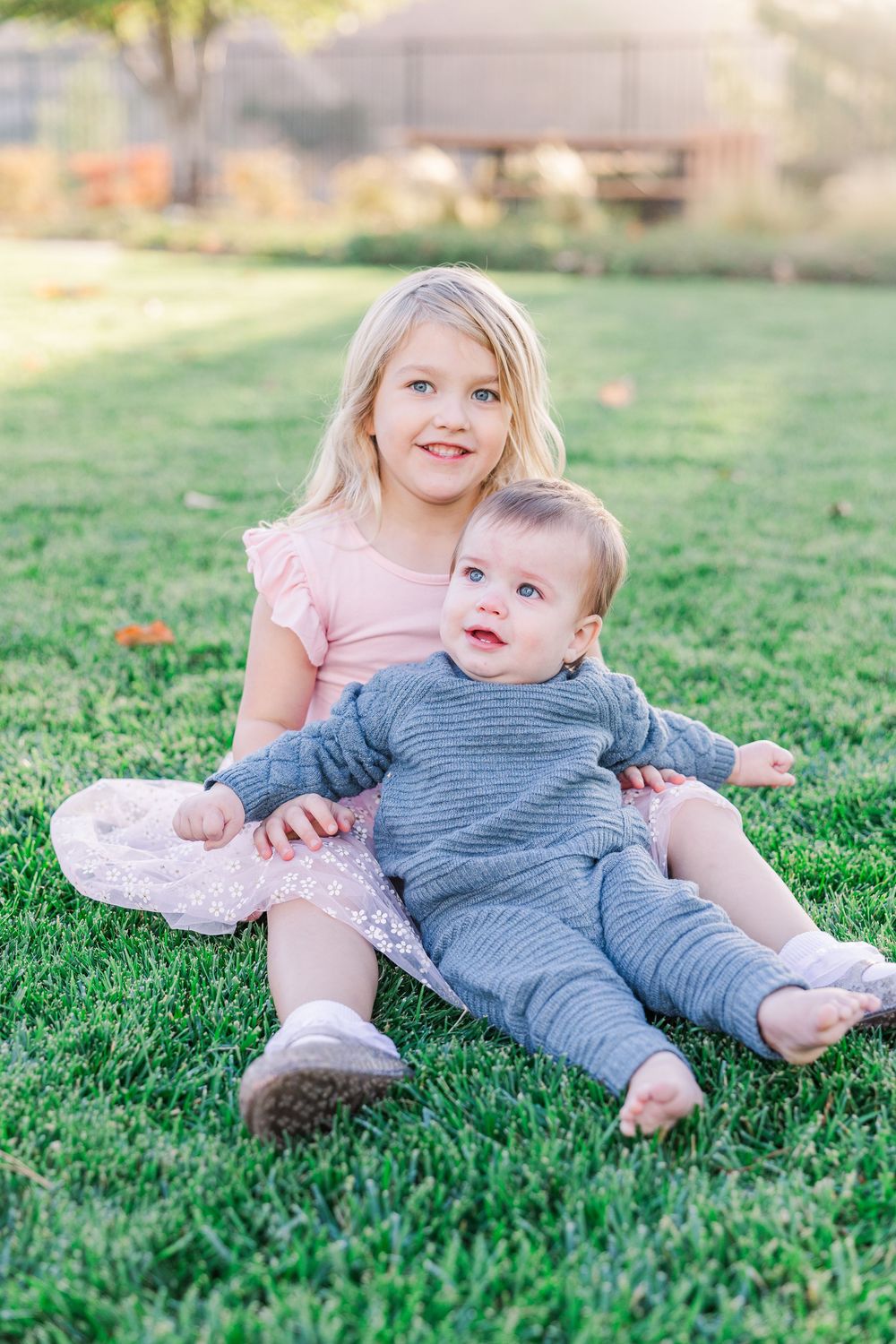 Two children in casual wear lying together on green grass during a sunny outdoor photoshoot.