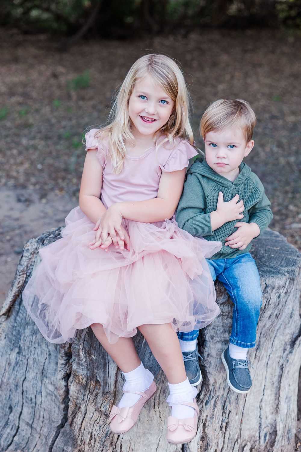 Two children sit together on a rustic wooden log, one wearing a pink dress and the other in casual clothing.