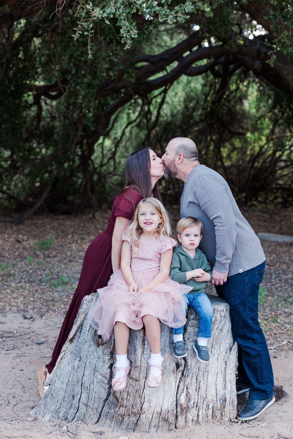 A family sits together on a large tree stump in a wooded area, sharing sweet moments in matching casual outfits.