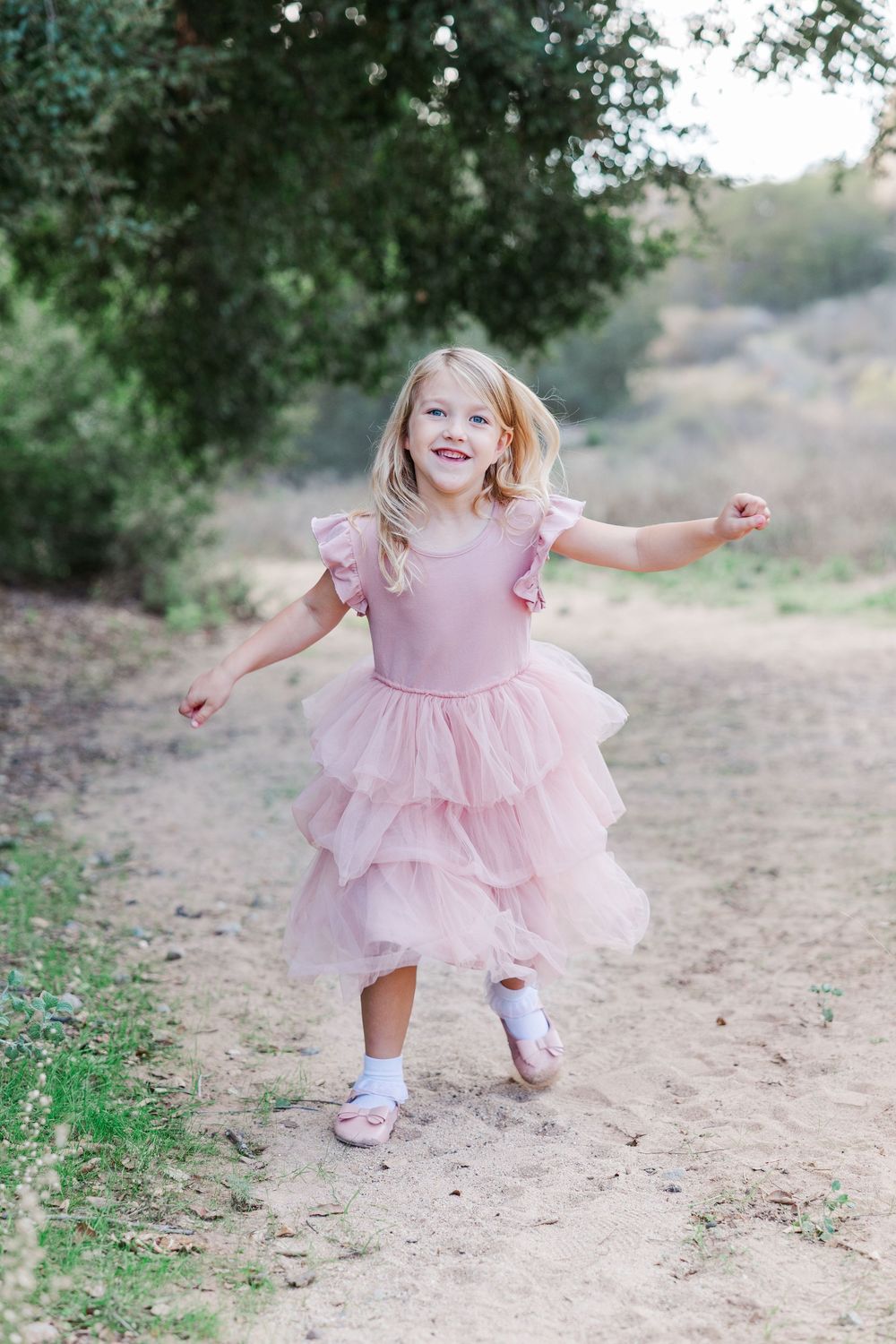 A child in a pink ruffled dress twirls and dances joyfully on a dirt path surrounded by greenery.