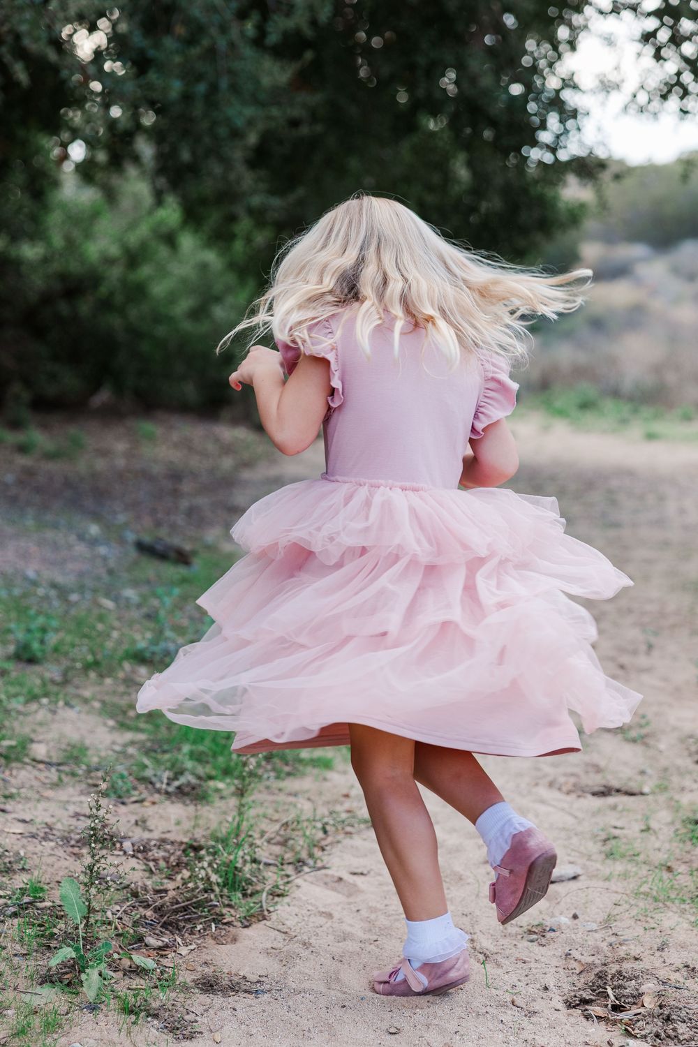 A pink twirling dress flows in motion as small feet in white socks and purple shoes dance on a dirt path.