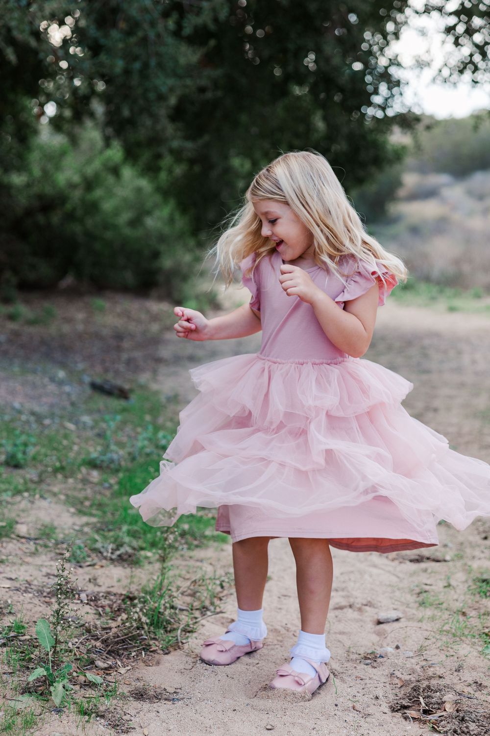 A young child twirls in a pink ruffled dress and sneakers on a dirt path surrounded by greenery.