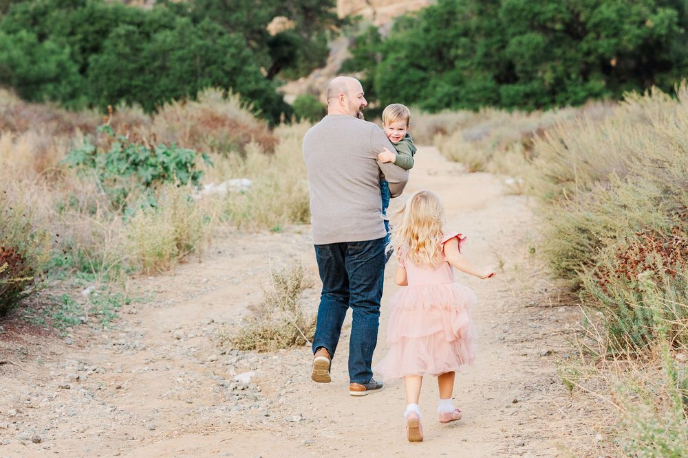People walk along a winding dirt path through dry grass and desert brush on a sunny day.
