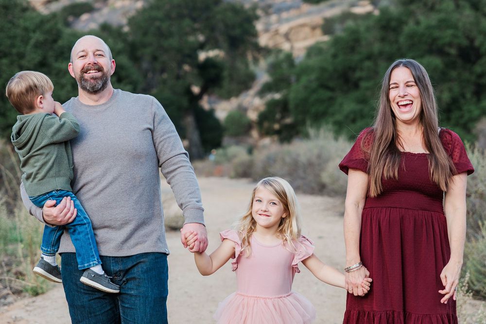 A family in casual clothing walks together and holds hands while smiling on a trail with hills in the background.