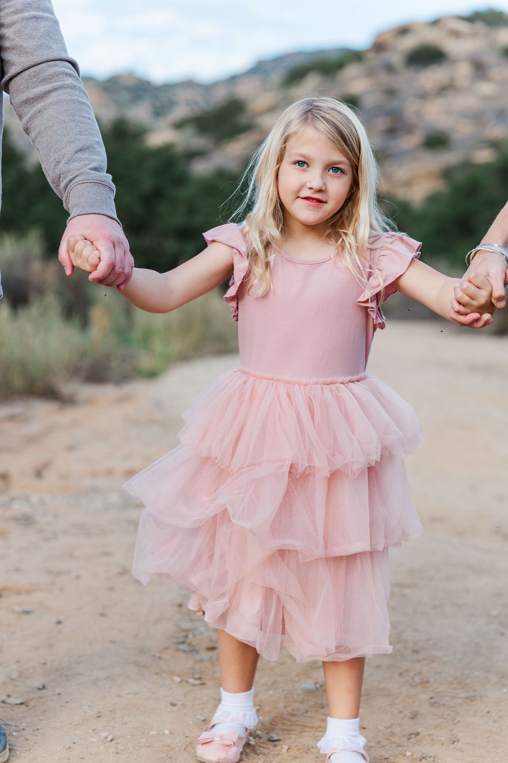 A small child in a pink tulle dress twirls and dances on a dirt path against a mountainous backdrop.