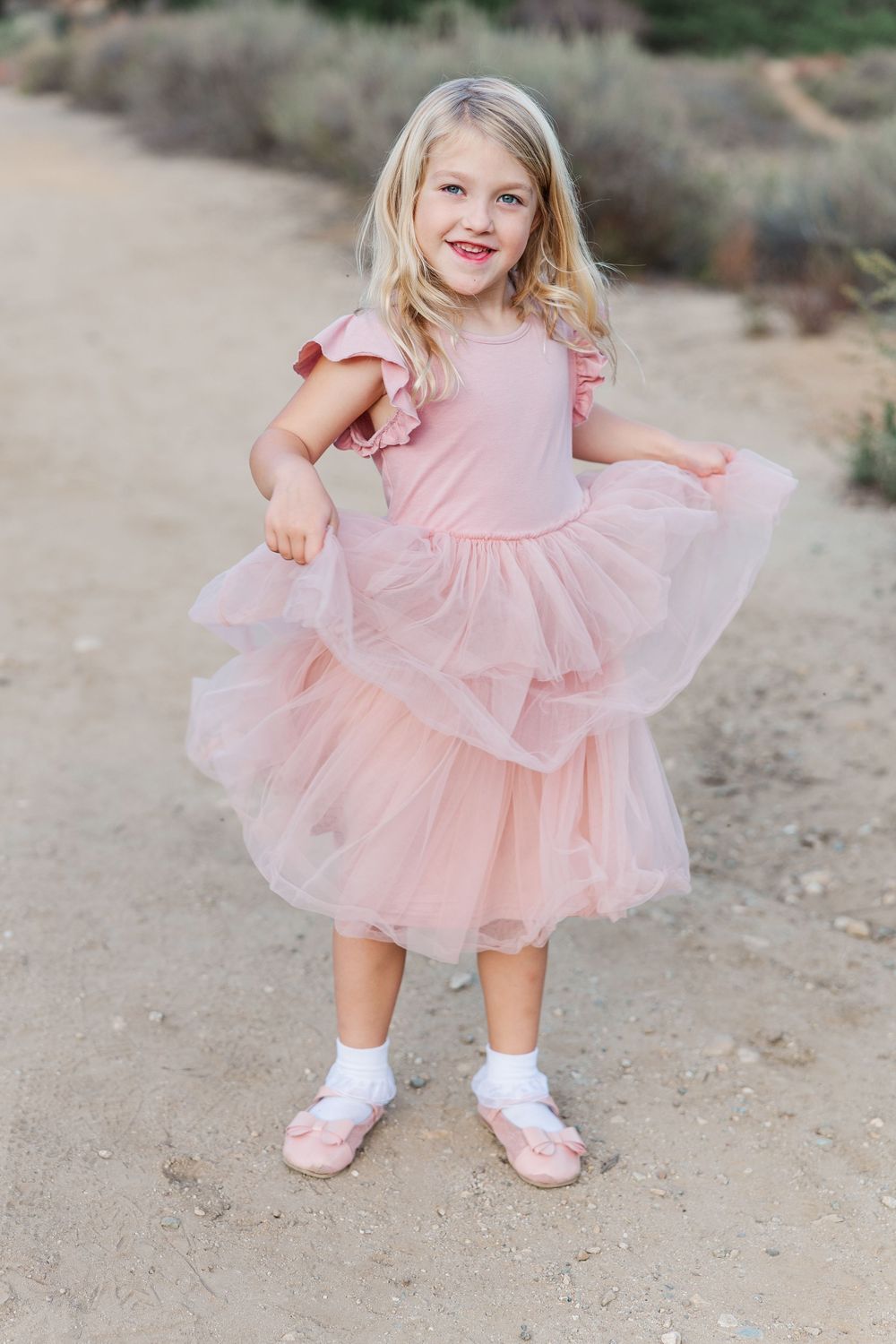 A child in a pink tulle dress twirls playfully on a dirt path outdoors.