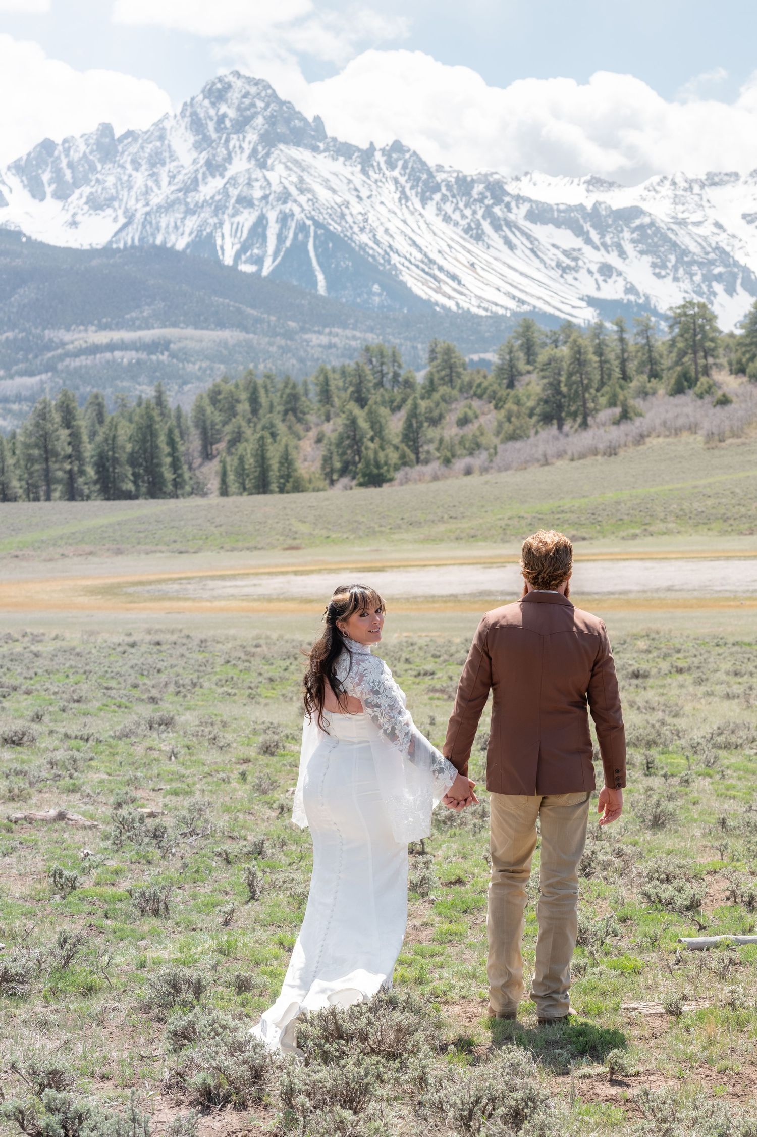 A bride and groom walk holding hands in a meadow. The San Juan mountains are snow covered in the background She looks back at the camera.