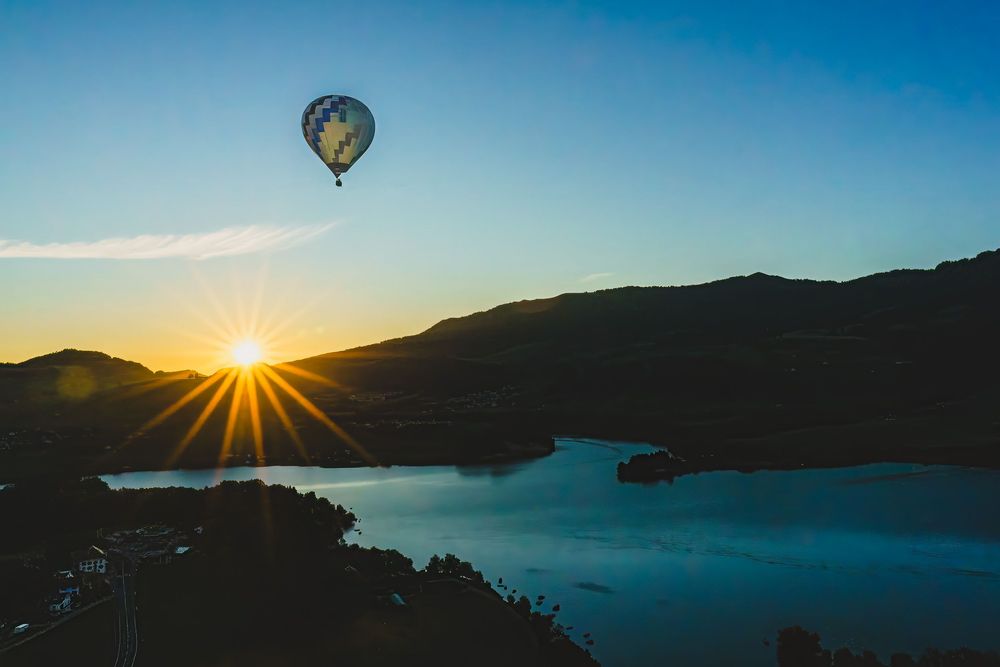 Hot air balloon floating over a serene mountain lake at golden sunset with vibrant sun rays and silhouetted hills.