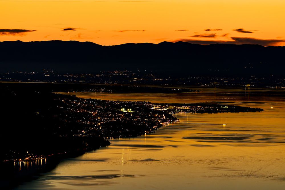 Aerial view of a shoreline at sunset with golden water reflections and distant mountains silhouetted against an orange sky.