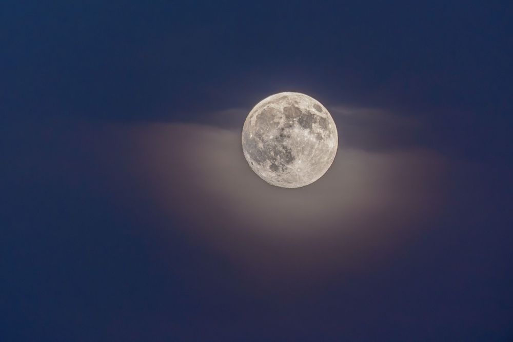 A bright full moon glows in a dark blue night sky with wispy purple clouds drifting across its illuminated surface.
