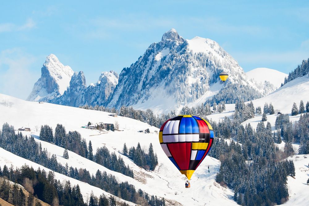 Colorful hot air balloon floating over snow-covered alpine mountains with evergreen trees on a clear winter day.