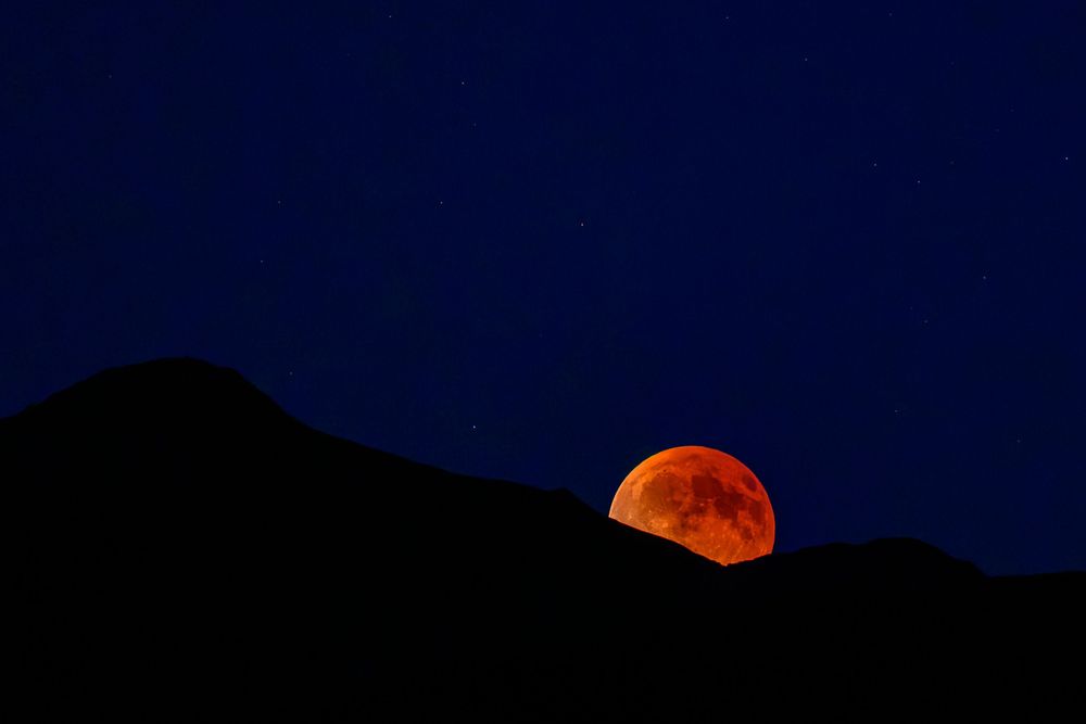 A vivid blood moon rising over dark mountain silhouettes against a deep blue night sky.