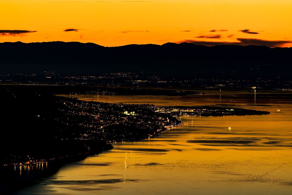 Aerial view of a coastal city glowing with lights at golden sunset, reflecting on calm water with mountain silhouettes.