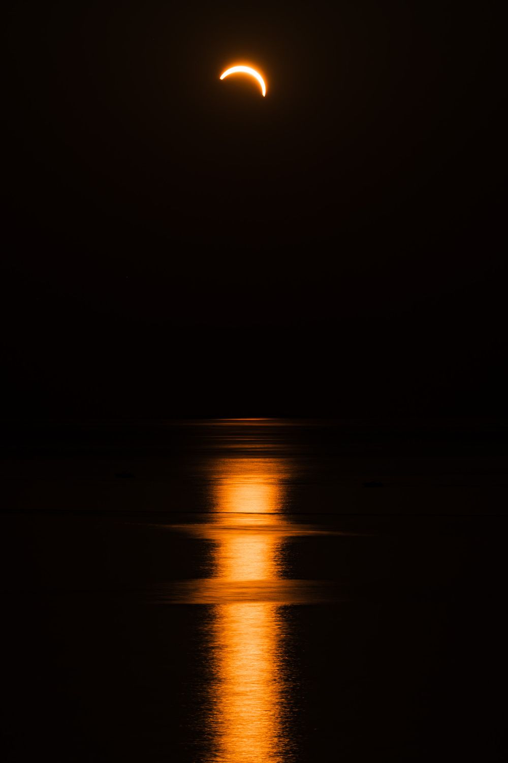 Partial solar eclipse reflected over dark ocean waters, glowing orange crescent against black sky.