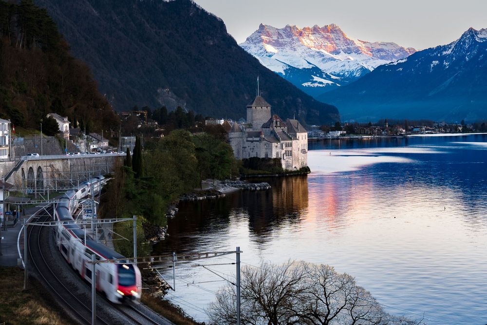 A modern train passes Chillon Castle on Lake Geneva with snow-capped Alps reflecting in calm waters at dusk.
