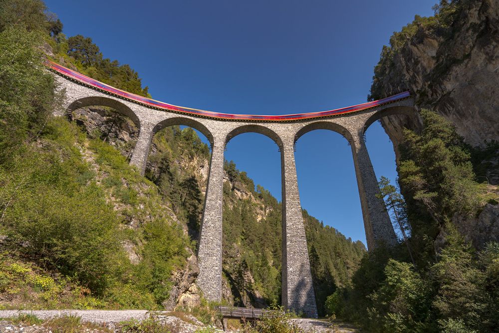 Red train crosses the iconic Landwasser Viaduct stone arches in Swiss Alps under clear blue sky.