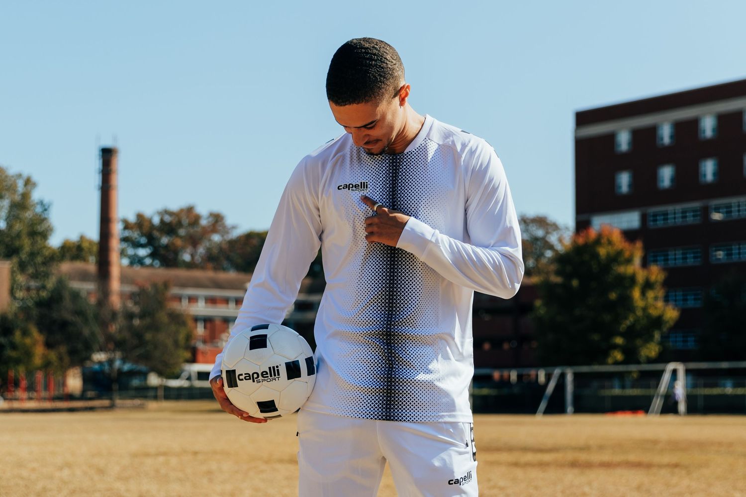 Male athlete in white Capelli Sport tracksuit holding a soccer ball on a sunny field.
