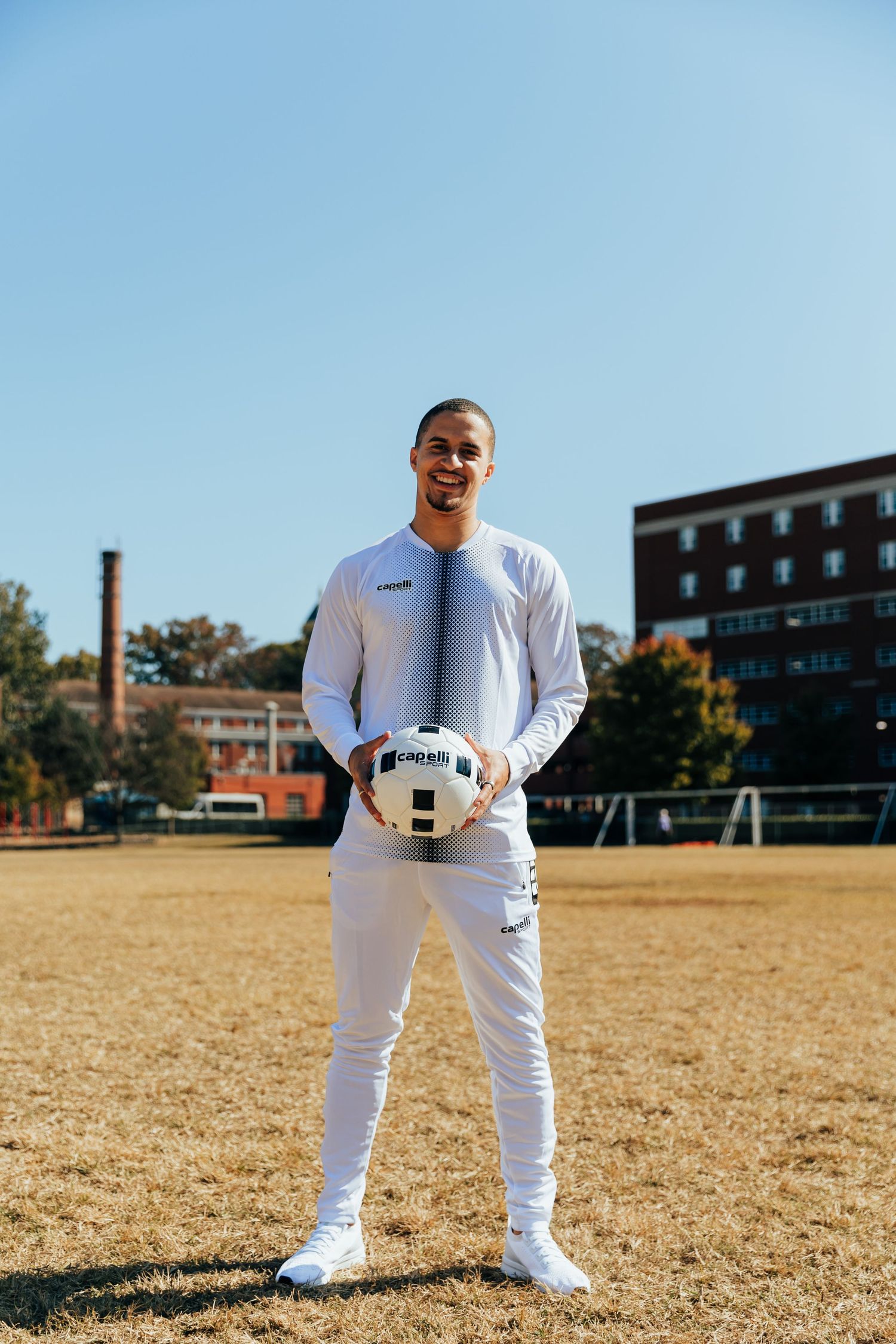 Young male soccer player holding a ball on an outdoor field wearing a white athletic outfit.